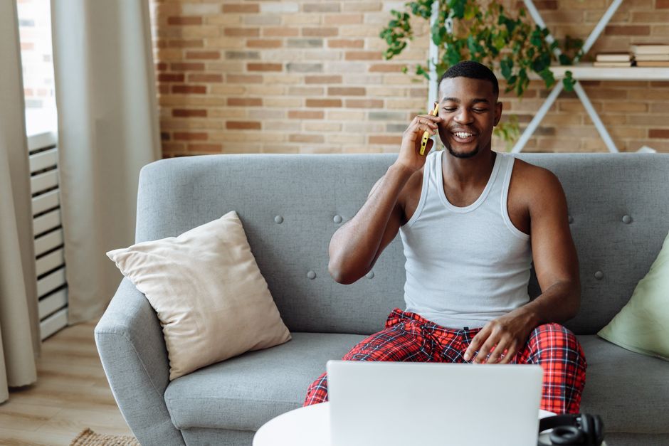 Homem relaxado conversando no celular enquanto está sentado no sofá, usando o laptop em um ambiente doméstico confortável.