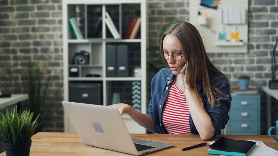 Mulher jovem e focada trabalhando no laptop enquanto conversa ao telefone em um escritório moderno e estiloso.