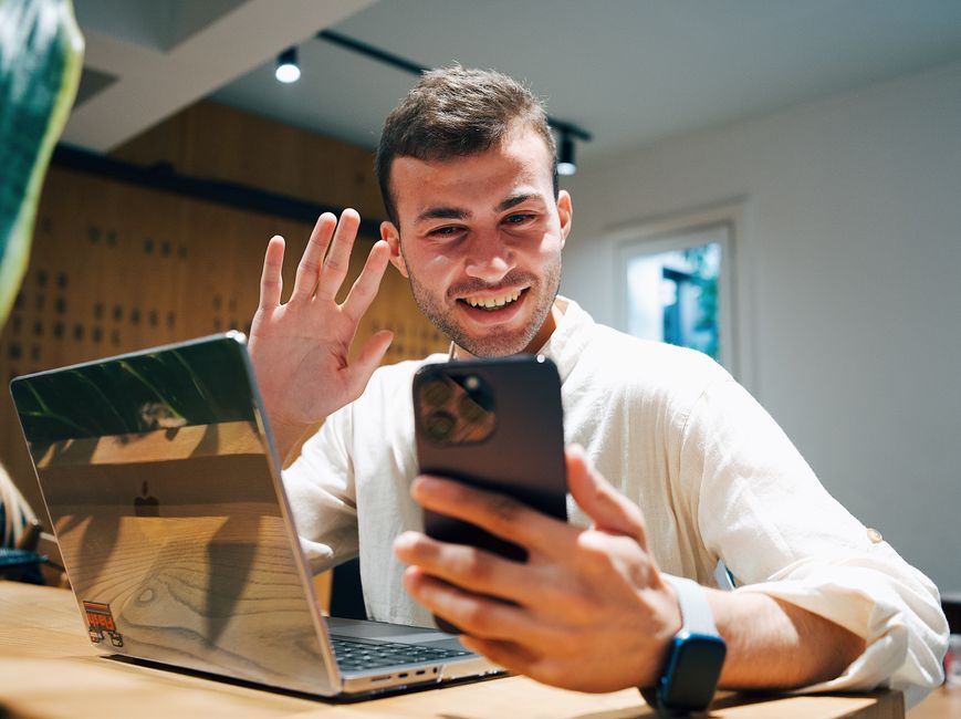 Jovem sorridente fazendo uma videochamada pelo smartphone enquanto está sentado com um laptop em um escritório moderno.