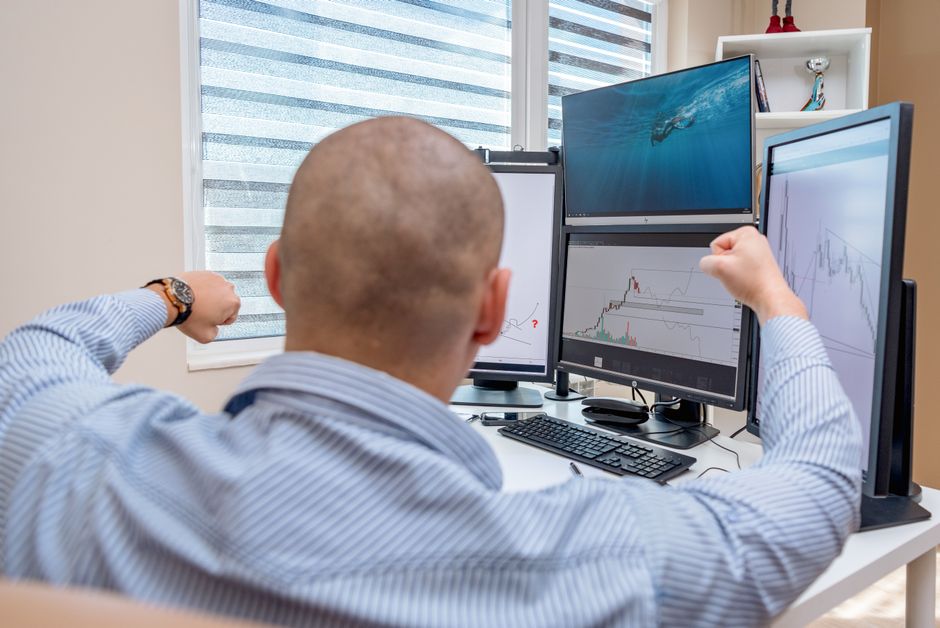 Um homem celebra o sucesso em uma estação de trabalho com múltiplos monitores enquanto analisa gráficos de ações.